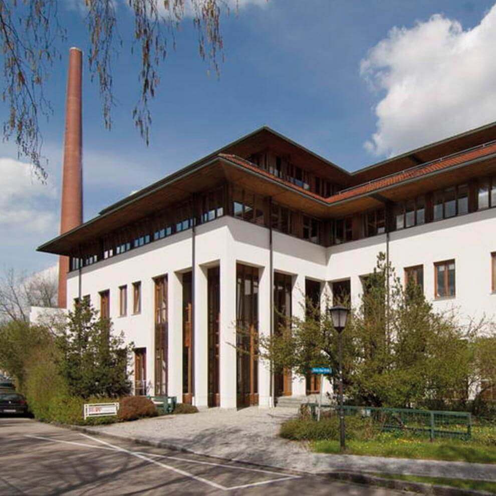 A modern building with a tall chimney stands prominently against a clear sky, surrounded by trees and shrubs.