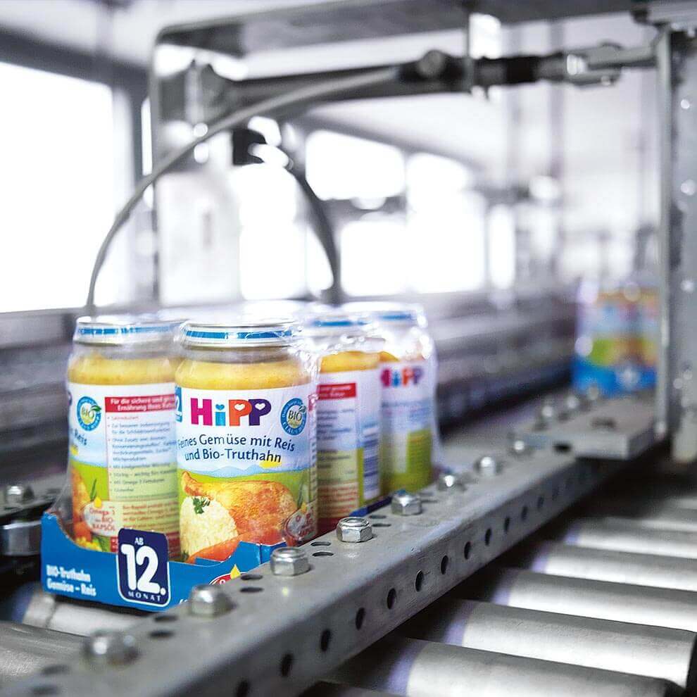 Jars of baby food with the label "HiPP" are moving along a conveyor belt in a factory setting.