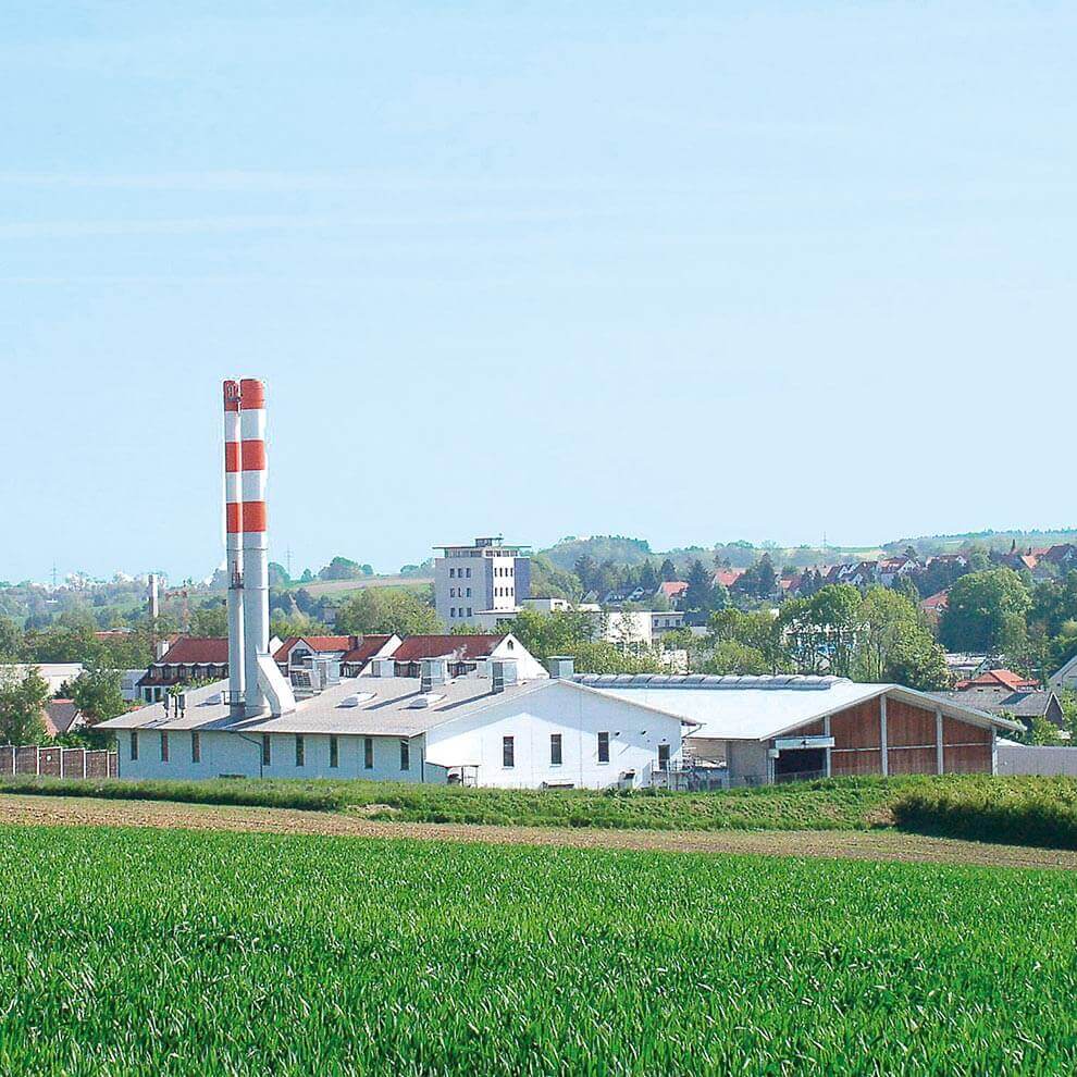 A factory with a tall chimney stands in the foreground, overlooking a town with various buildings and a church steeple in the distance, all set against a clear sky.