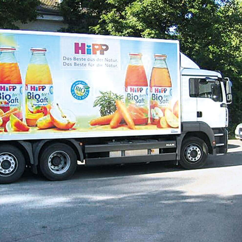 A white truck is parked on a street, displaying a large advertisement for Hipp Biosaft beverages, which are depicted as colorful and fresh fruit and vegetable juices.