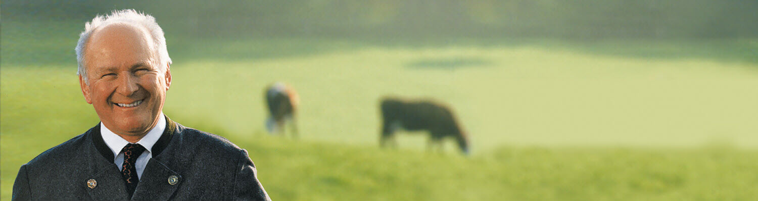 An elderly man in a suit stands in a field with cows grazing in the background.